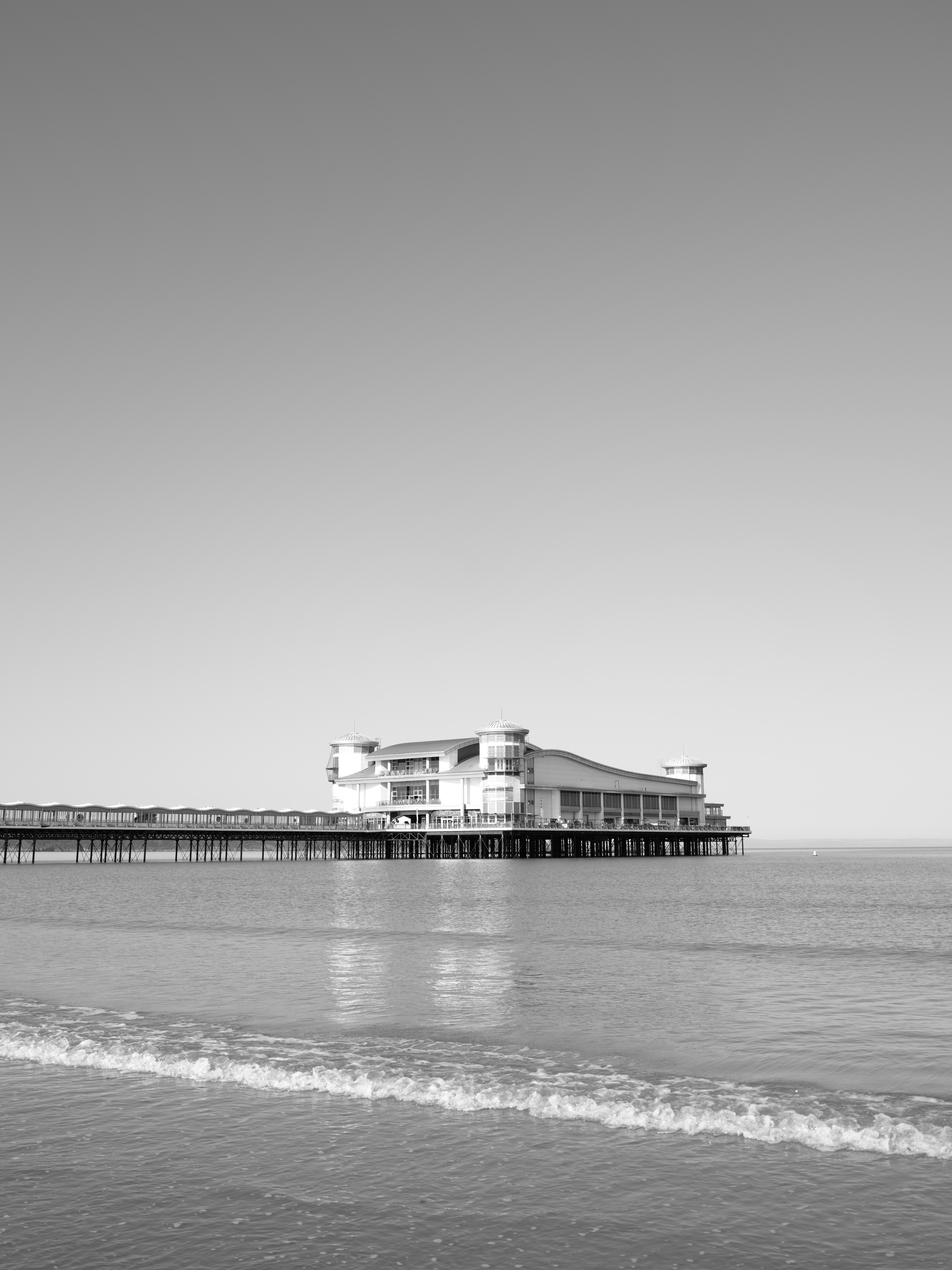 Black and white image of a seaside pier with a large building atop, reflecting on calm water under a clear sky.