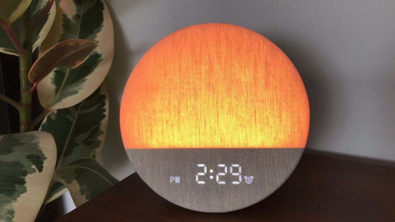 A round sunrise alarm clock emitting a soft glow light next to a green plant in a sleep tester's bedroom