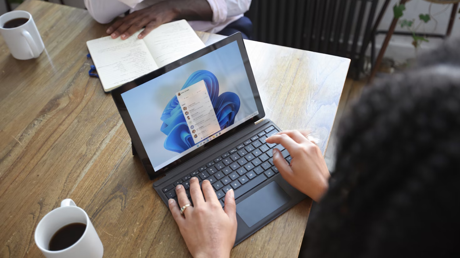 Una mujer en una mesa usando una laptop con Windows 11, frente a ella un hombre, sin verse sus rostros