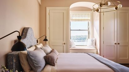 A calming bedroom painted a soft pink with cream built-in wardrobes and an arched window with a seating nook. A large bed with mauve bedding, blue cushions and two black wall lights.