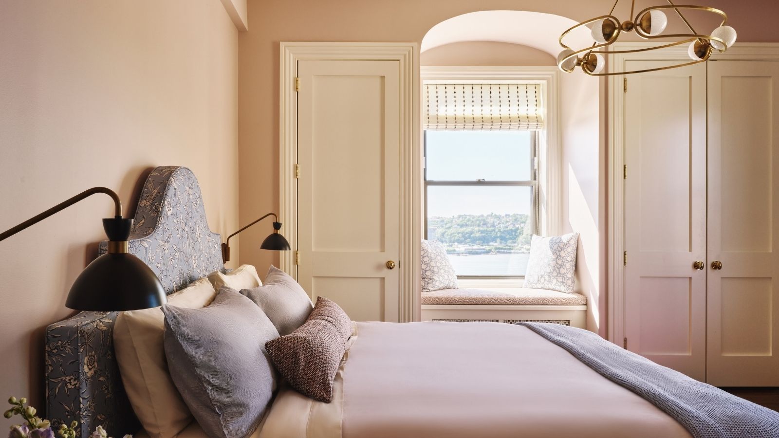 A calming bedroom painted a soft pink with cream built-in wardrobes and an arched window with a seating nook. A large bed with mauve bedding, blue cushions and two black wall lights.