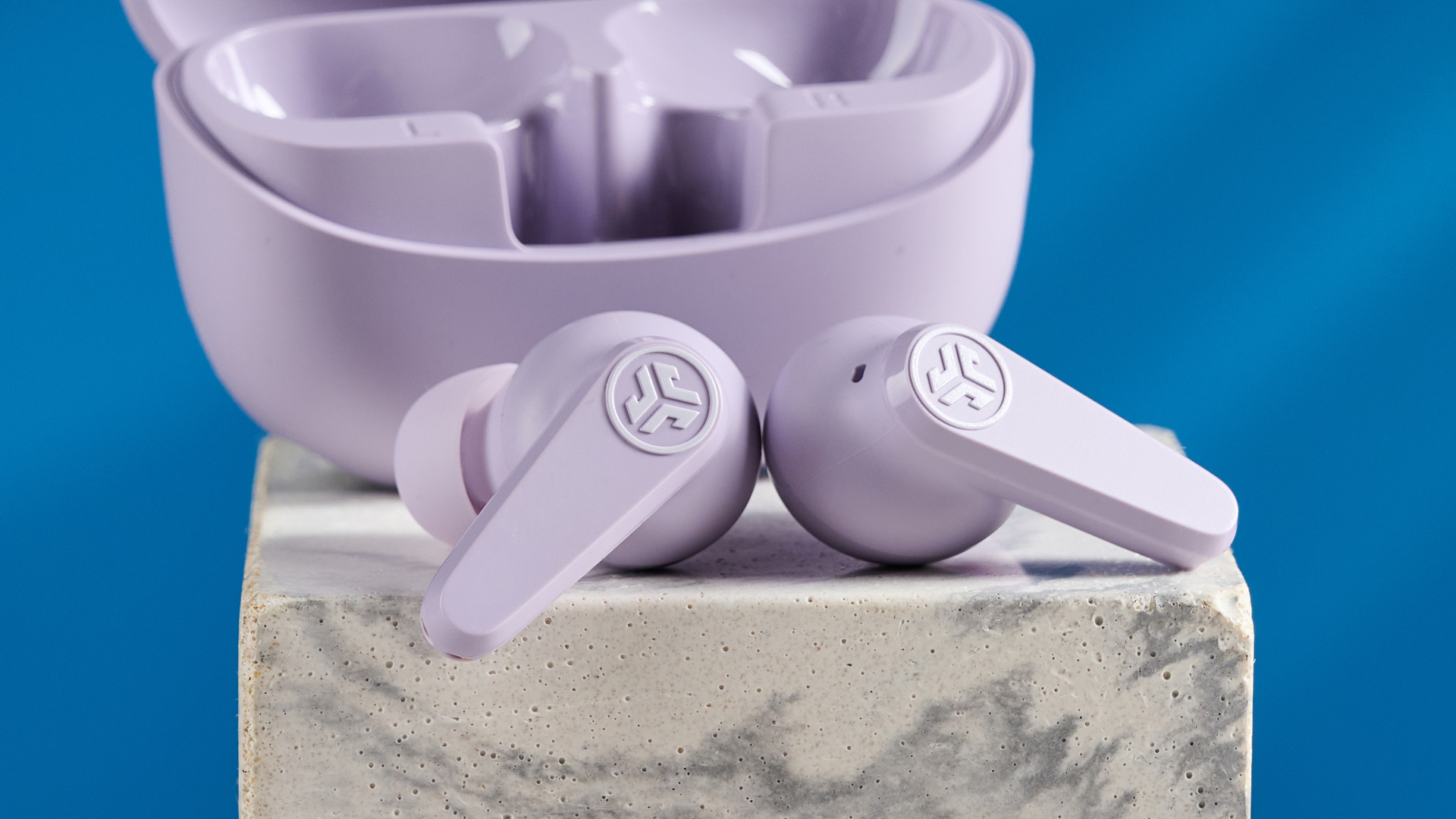 the jlab go pods anc earbuds in lilac with a lilac charging case and built in usb-c cable against a blue background on a marble stand