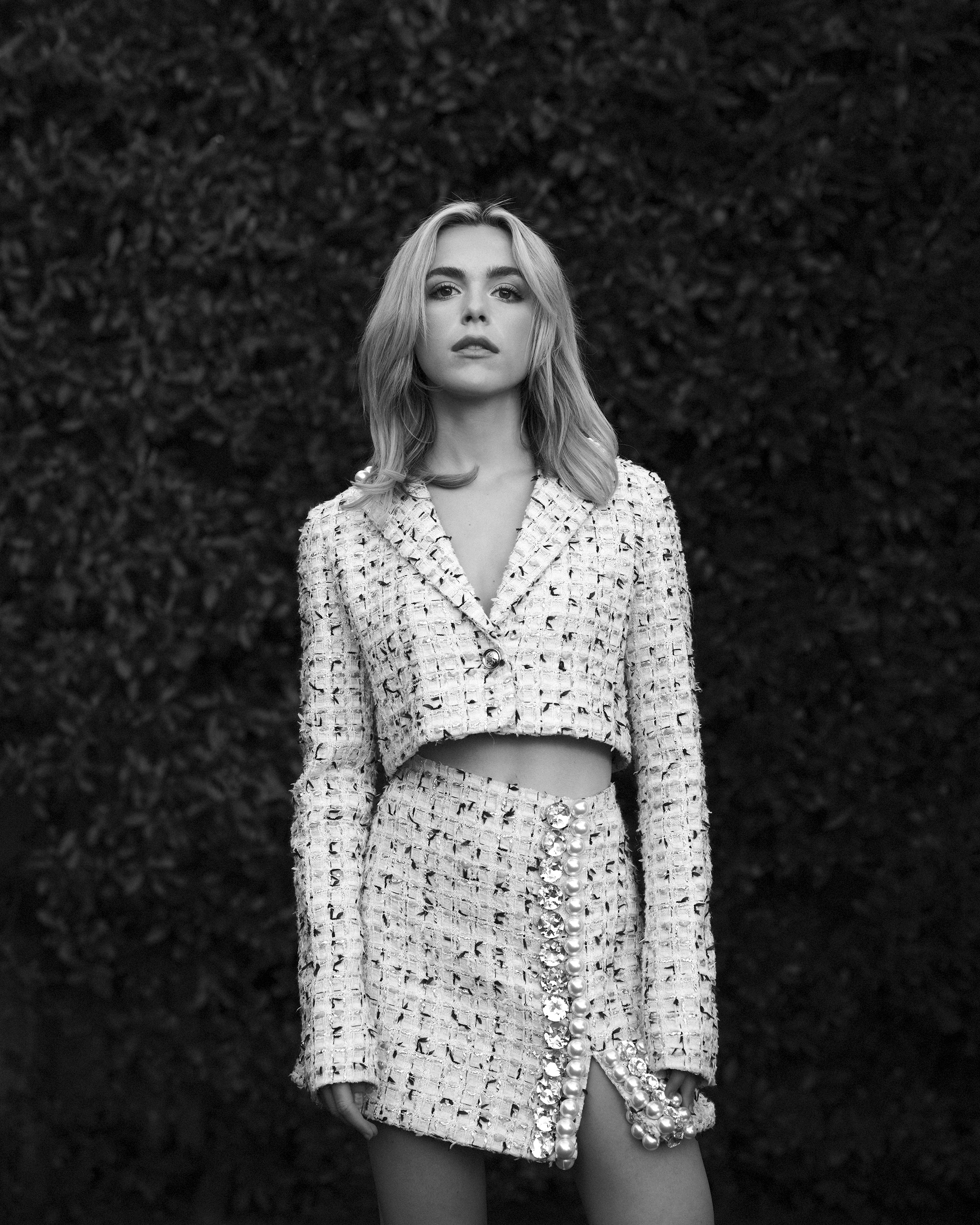 kiernan shipka wears a chanel two piece cropped suit poses in front of a hedge in a black and white photo