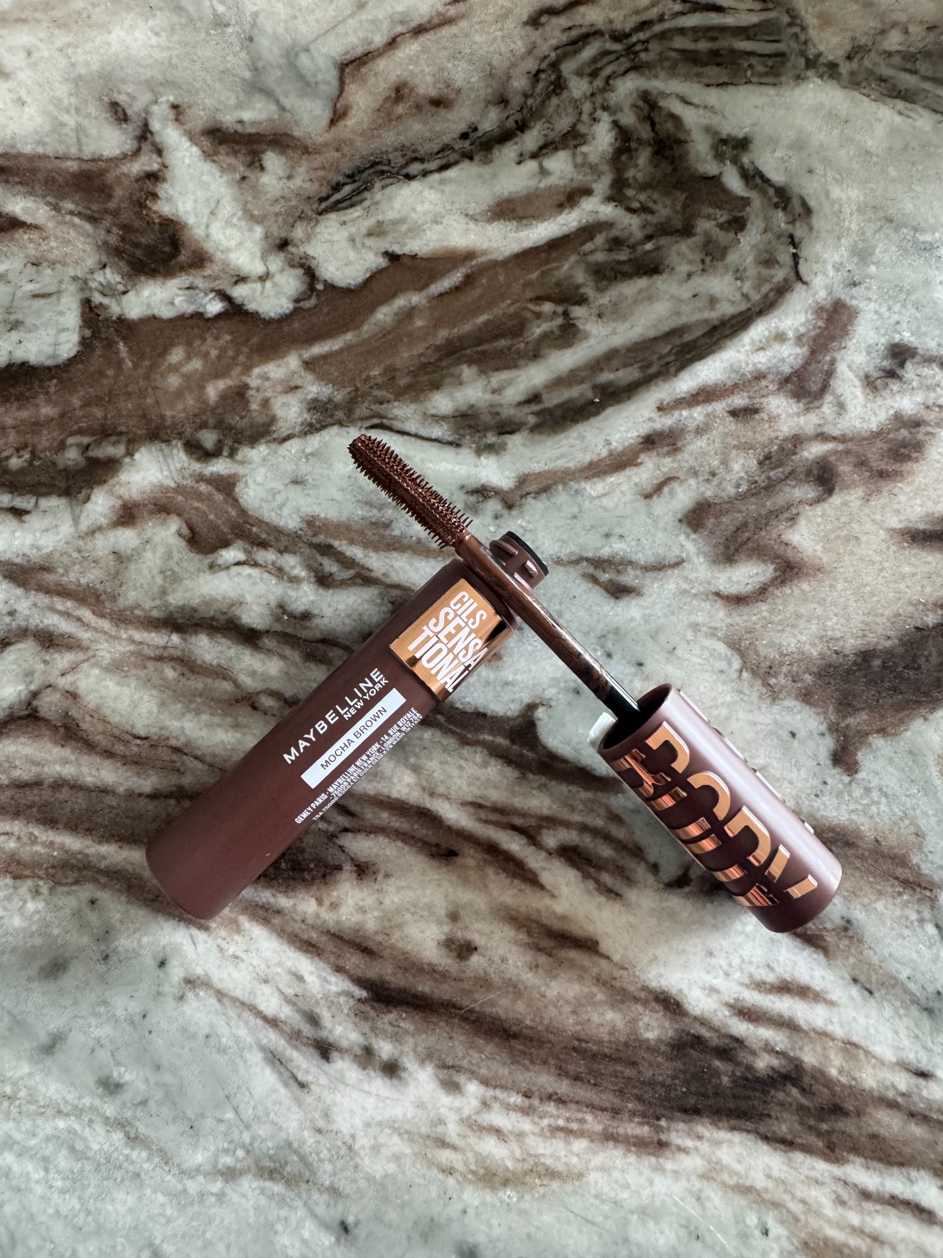The Maybelline Lash Sensational Body Mascara in Mocha brown shade on a marble table, with the wand out