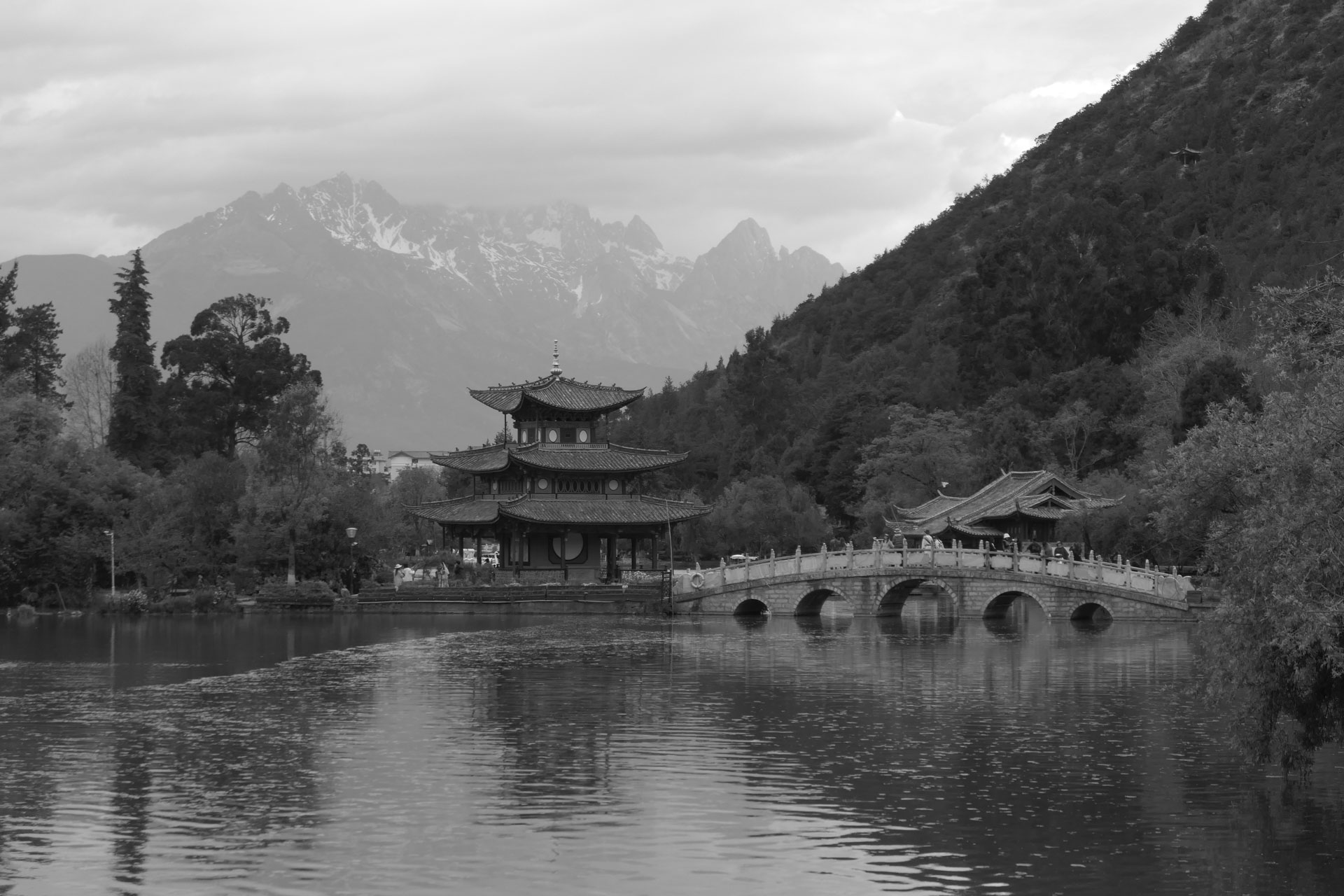 A Chinese temple by the side of a lake with an arched bridge leading up to it and both are reflected in the still water, in front of a snow covered mountain. A creative filter is applied to the image