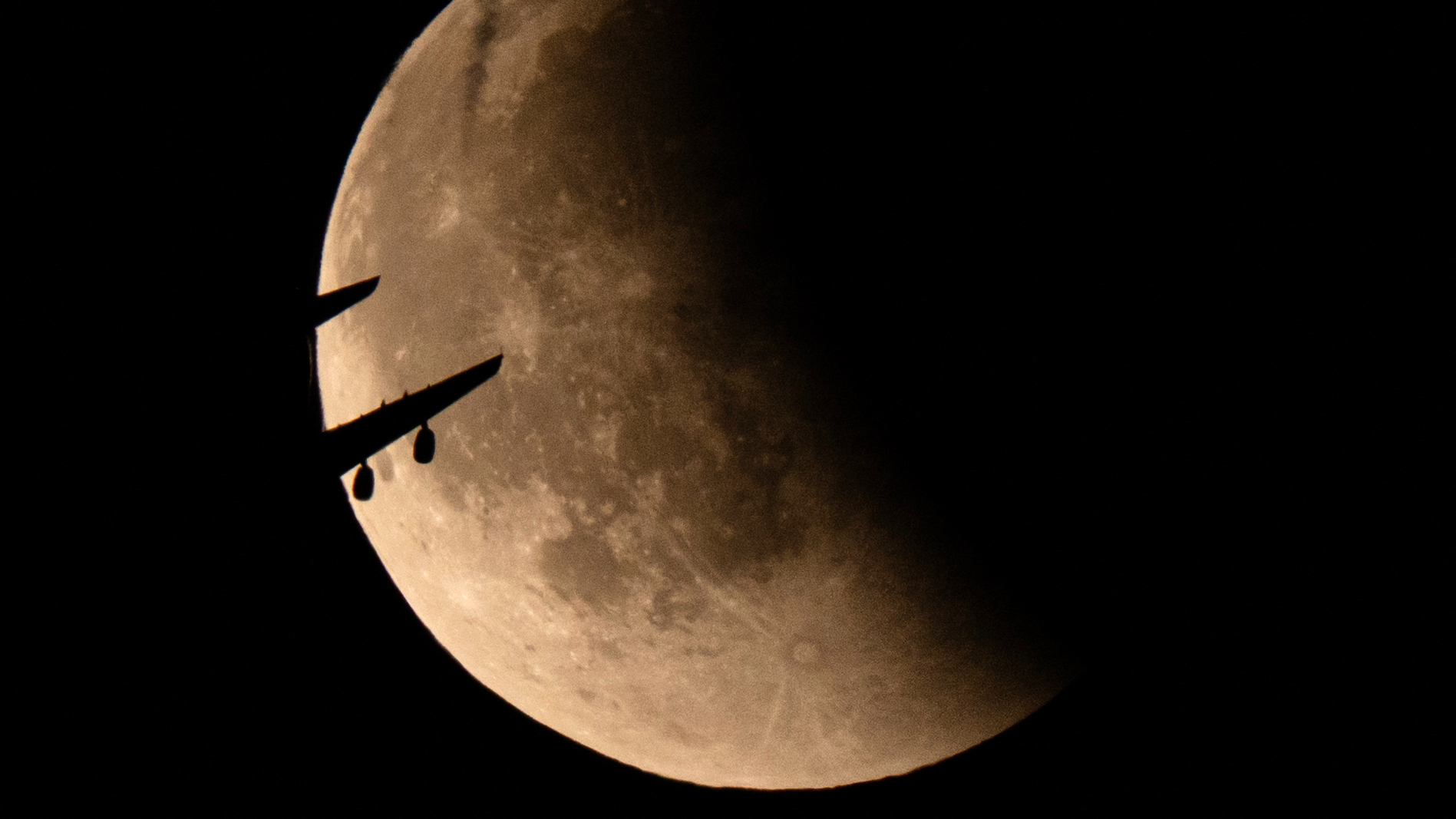 A full moon is shown against a black sky with its right half bathed in shadow during a lunar eclipse. The silhouette of a plane can be seen crossing the lunar disk on its left side.