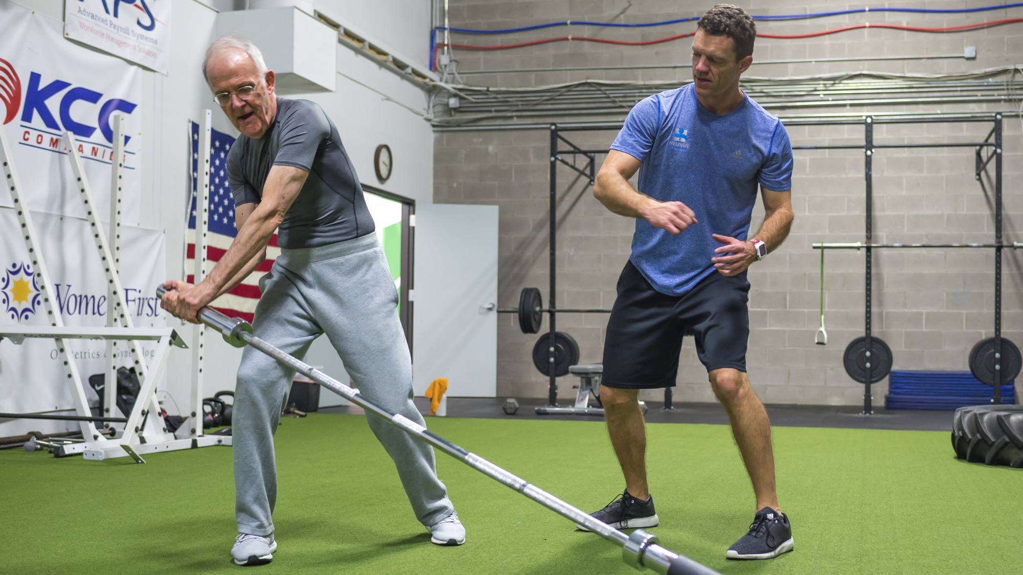 Senior man exercises with barbell, a personal trainer stands to the side mimicking the senior's movement 