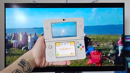 Hand holding up New 3DS with monitor in backdrop playing Artemis II 3DS launch footage by Cambot72.