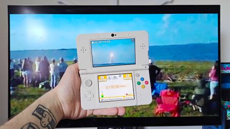 Hand holding up New 3DS with monitor in backdrop playing Artemis II 3DS launch footage by Cambot72.