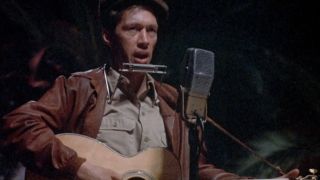 David Carradine with a guitar and a harmonica and singing into a microphone in Bound for Glory