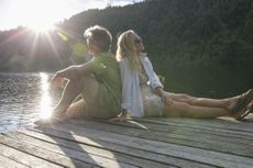 A mature couple in their late 40s or early 50s relax on lake dock in the sunshine.