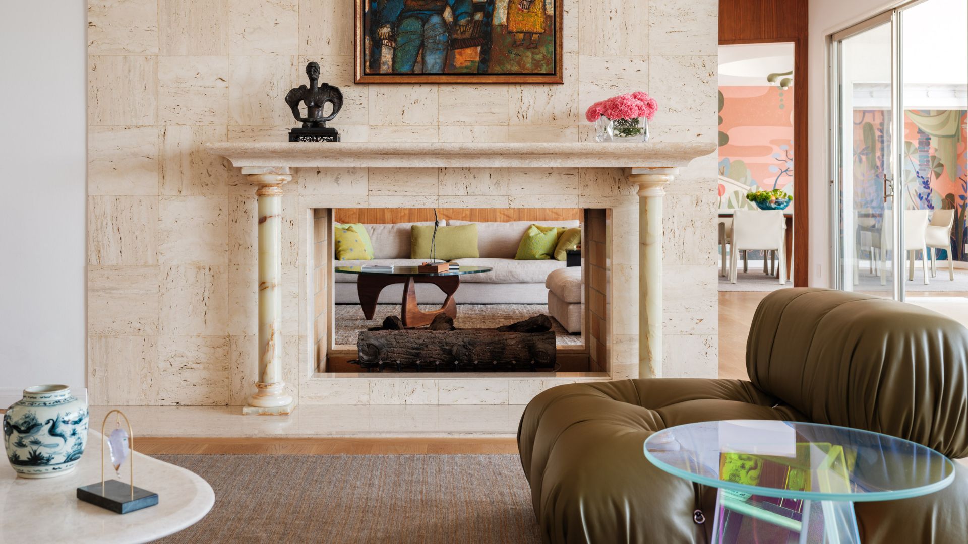A living room with an open fire place, a marble mantel, a green leather sofa seater, an iridescent side table, a vase of pink hydrangeas, a framed painting, a gray area rug and a side table with a ceramic jar