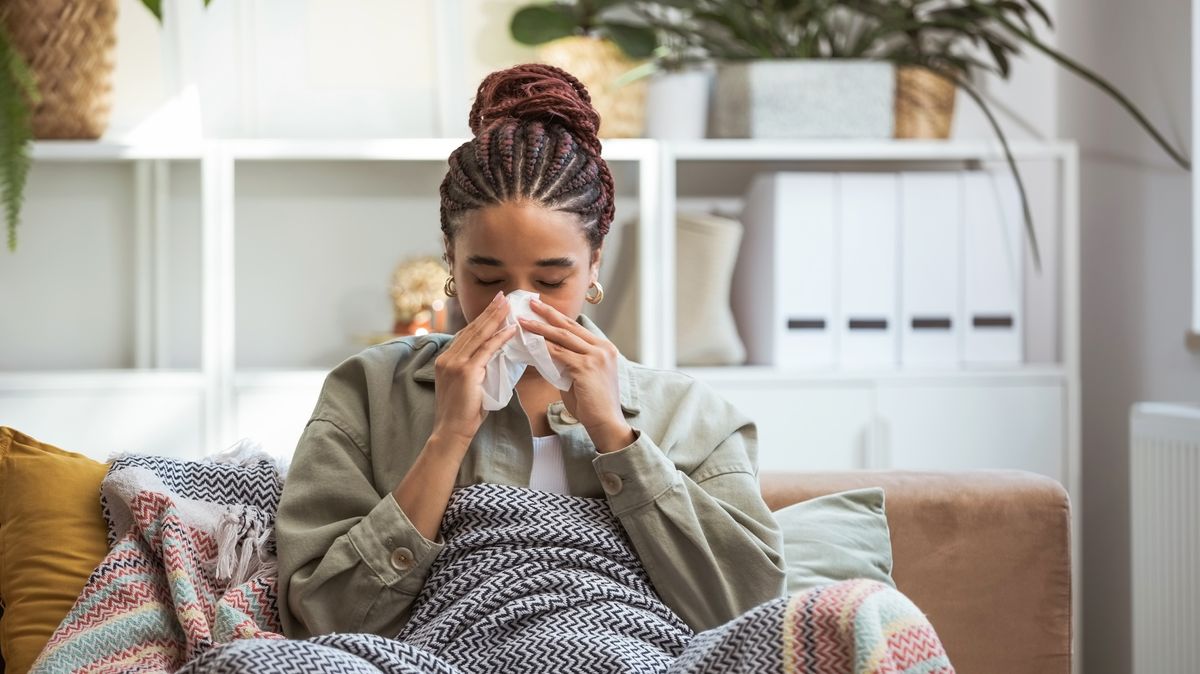 A woman sits on the couch under a blanket and blows her nose