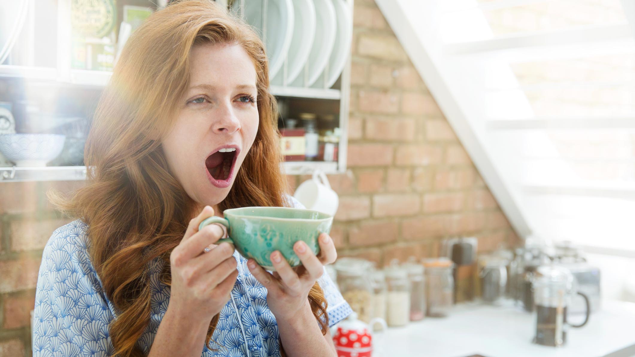 A woman with red hair stands in her kitchen yawning while holding a green tea cup