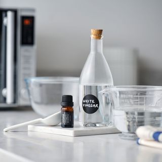 bottle of vinegar, bottle of essential oil, water jug on a worktop with a microwave in the background