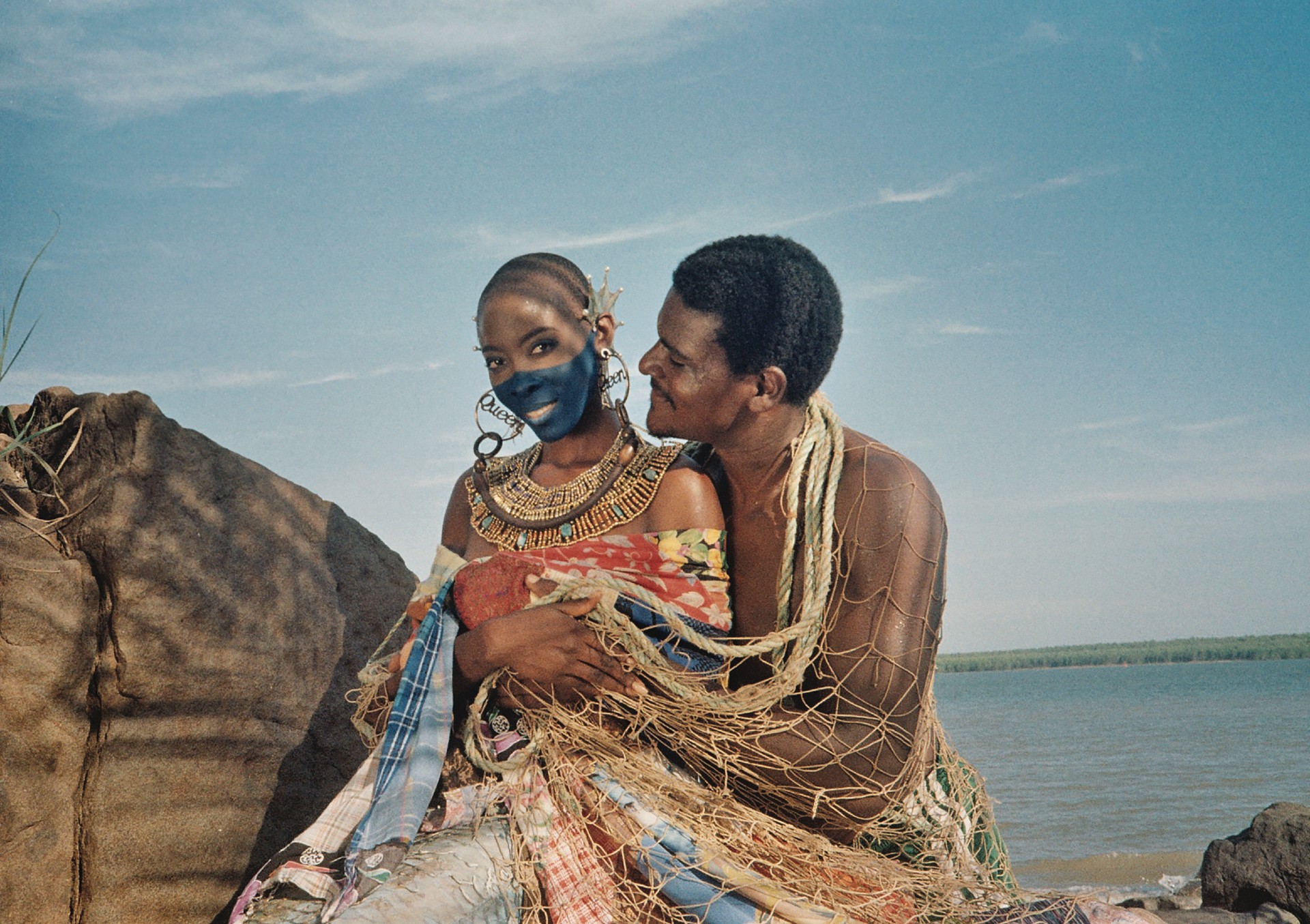 A man and a woman in ornate, colorful attire and blue face paint pose together on a rocky shore, partially draped in a large fishing net against a blue sky.