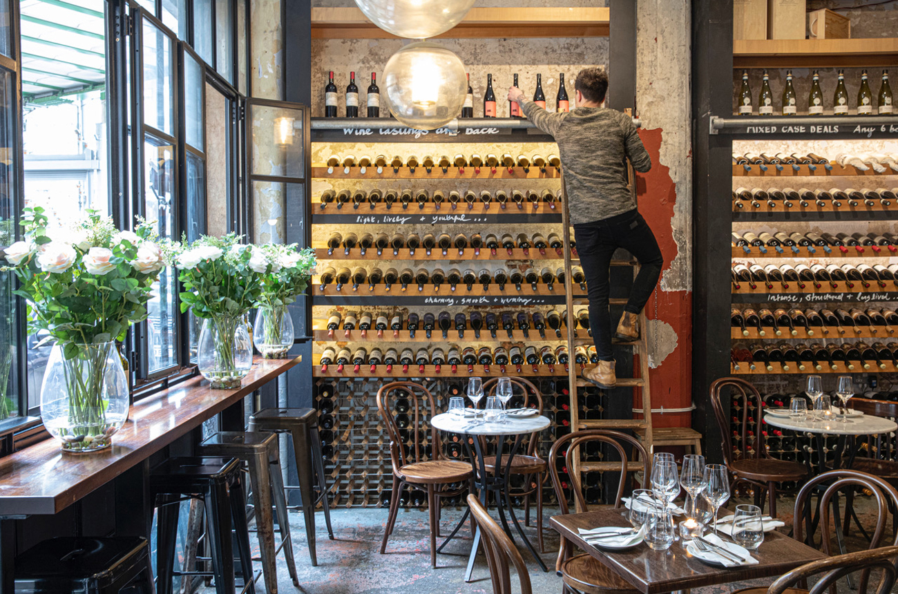 A man on a ladder in front of a wall of wine bottles in a wine bar
