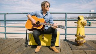 Chris Shiflett, posing with an acoustic guitar against a beach backdrop