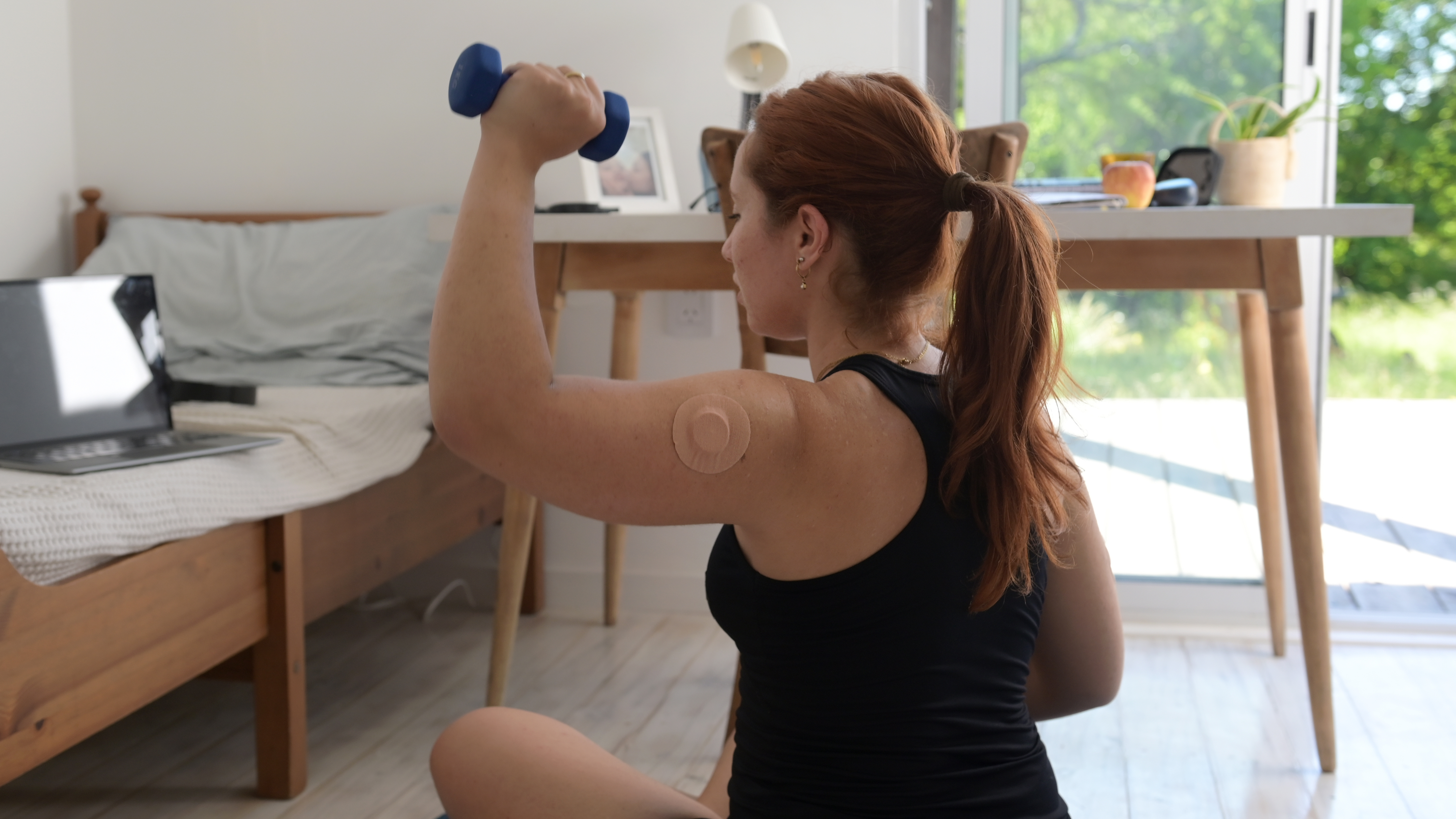 A woman with a glucose monitor on her arm holds a light dumbbell in her hand. She is sitting, with the dumbbell held at head height.