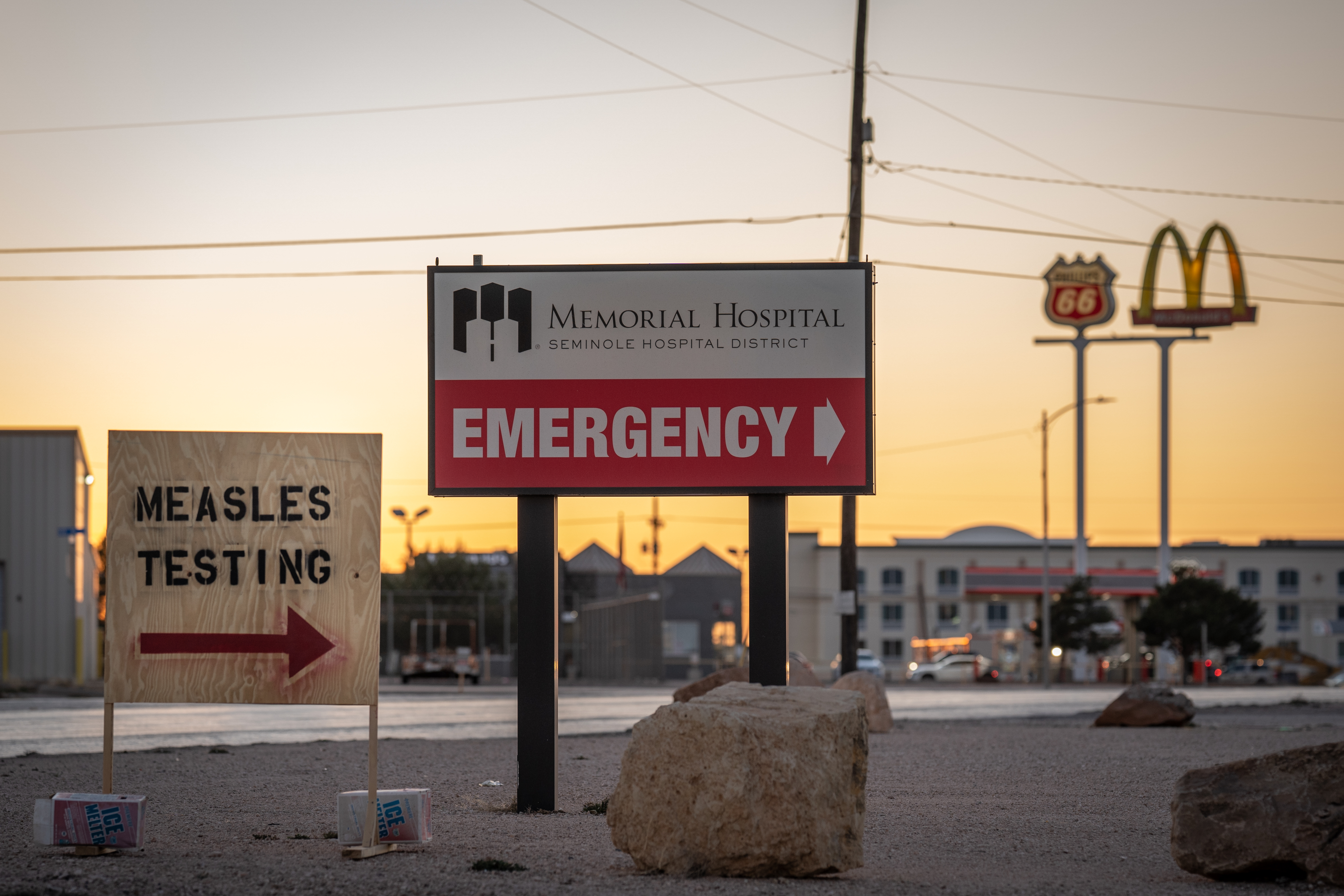 an emergency hospital sign at sunset with a board saying measles testing and an arrow, and macdonalds in the background