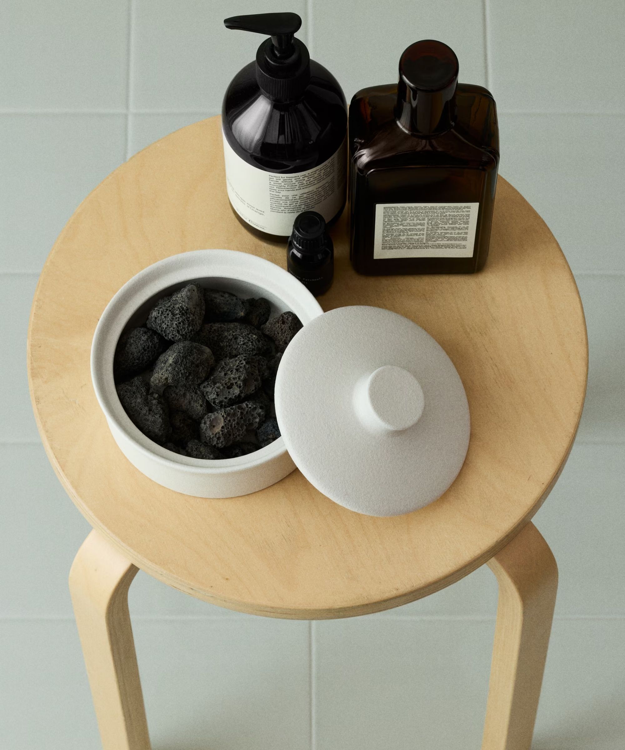 White H&amp;amp;M Lava Stone Diffuser filled with black lava stones shown on a pale wood stool with two black bottles against pale green tiles