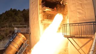 flames shoot out of a cone-shaped rocket engine poking out of a hangar-like building