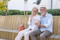 An elderly couple sits joyfully on a park bench, engaging with a tablet together in an outdoor setting, showcasing happiness, companionship, and the benefits of digital technology.