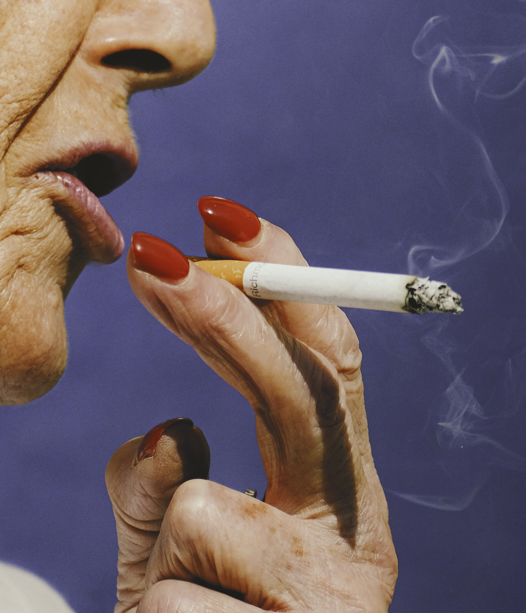 Close-up of an older woman's face exhaling smoke while holding a cigarette, with bright red nail polish on her fingers against a purple background