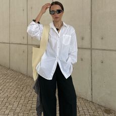 Debora wears a white button-down shirt, black pants, and an ivory leather shoulder bag.