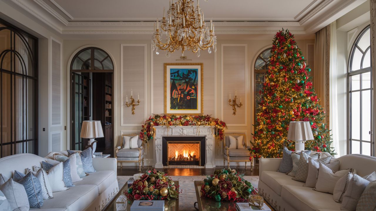 Ornate living room, featuring a crystal chandelier, a roaring fireplace, and oversized gray sofas, decorated for the holidays with a tall Christmas tree in the corner.