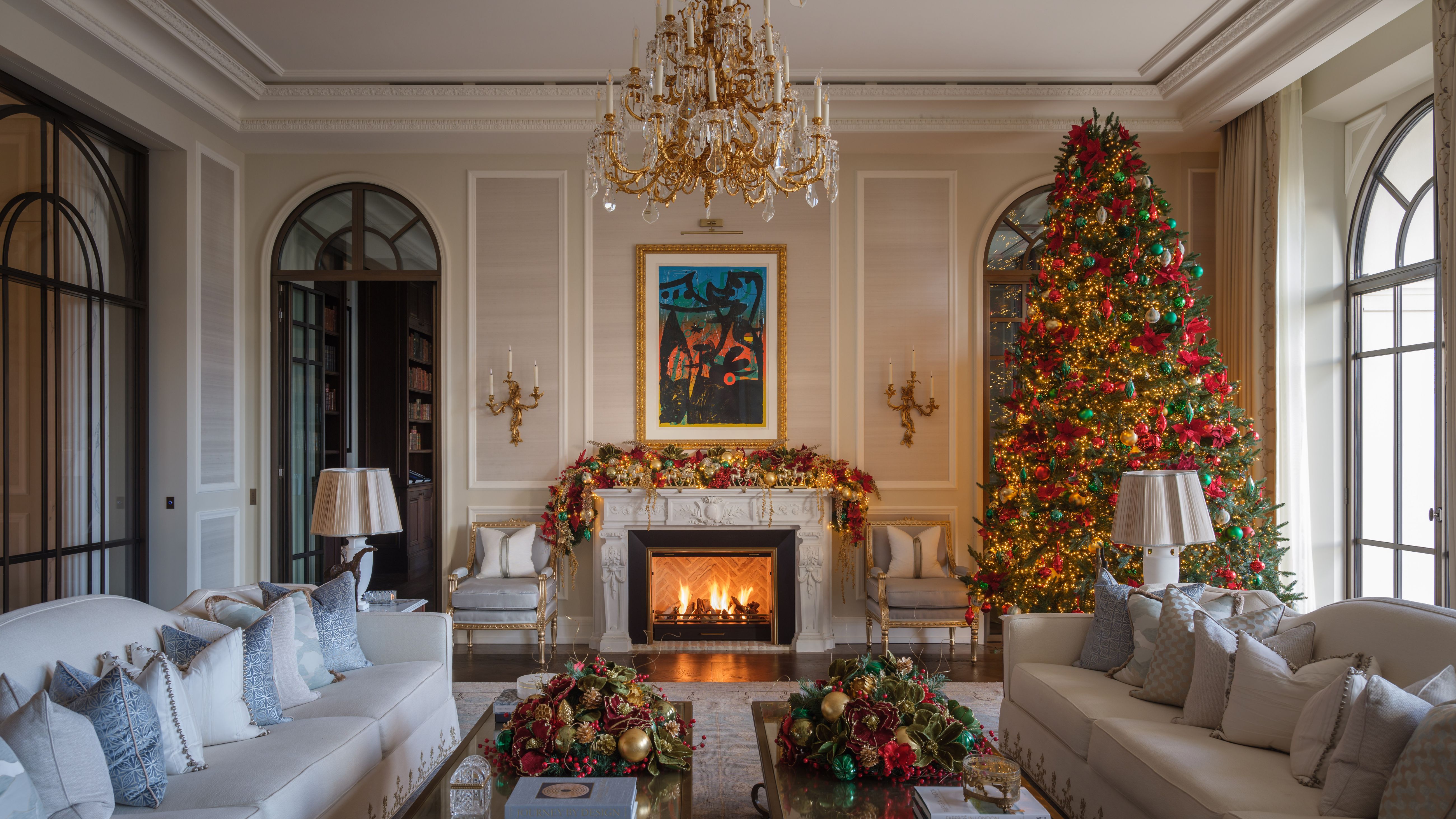 Ornate living room, featuring a crystal chandelier, a roaring fireplace, and oversized gray sofas, decorated for the holidays with a tall Christmas tree in the corner.