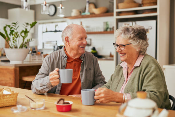 Smiling old couple sitting at table and sharing coffee in their cozy kitchen. Elder man and retired woman laughing together while enjoying a cup of coffee indoors.