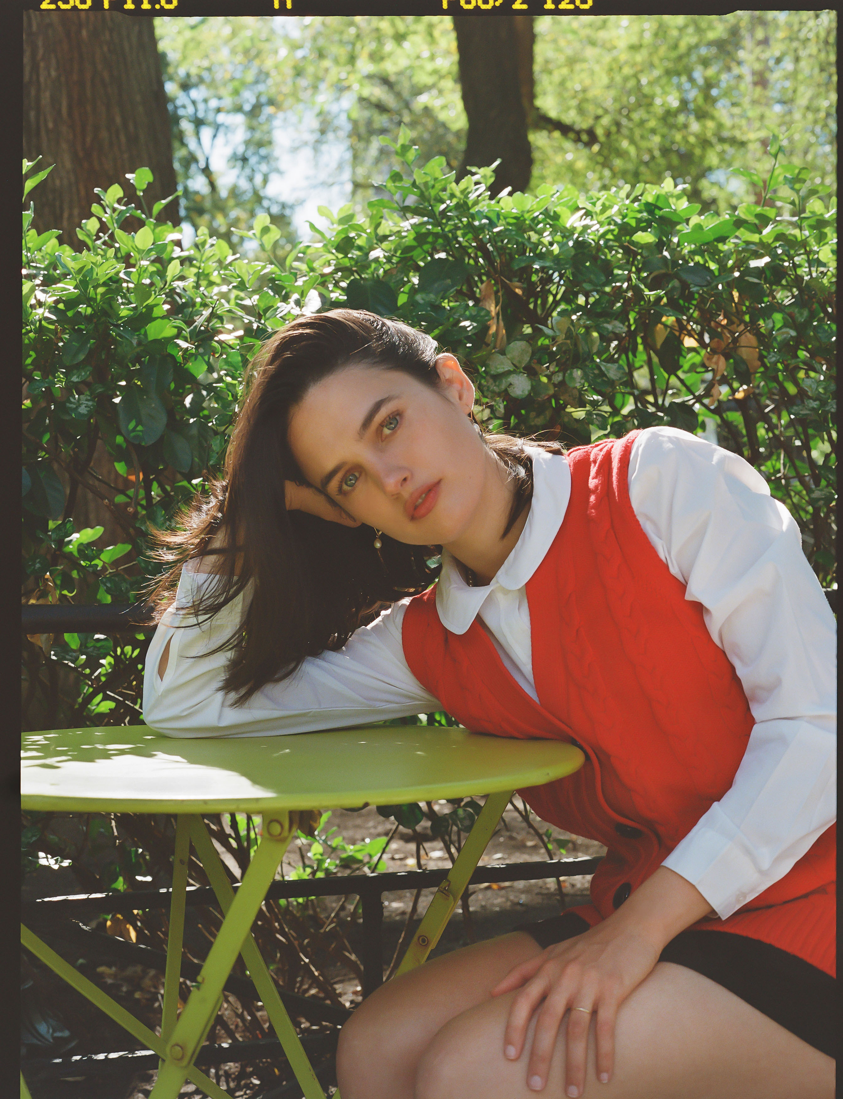 creative director nat ruiz from forever magazine poses on a park table by leaning on it and wears a red sweater vest over a white shirt with a peter pan collar