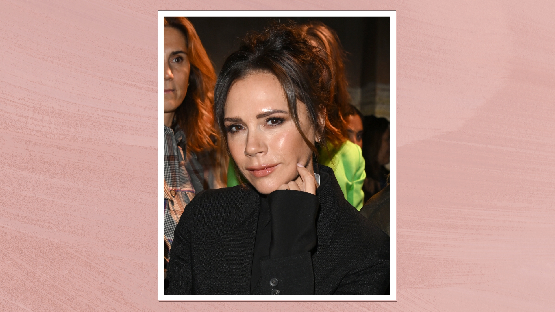 Image of Victoria Beckham smiling with brunette up-do and wearing a black blazer, on a pink background