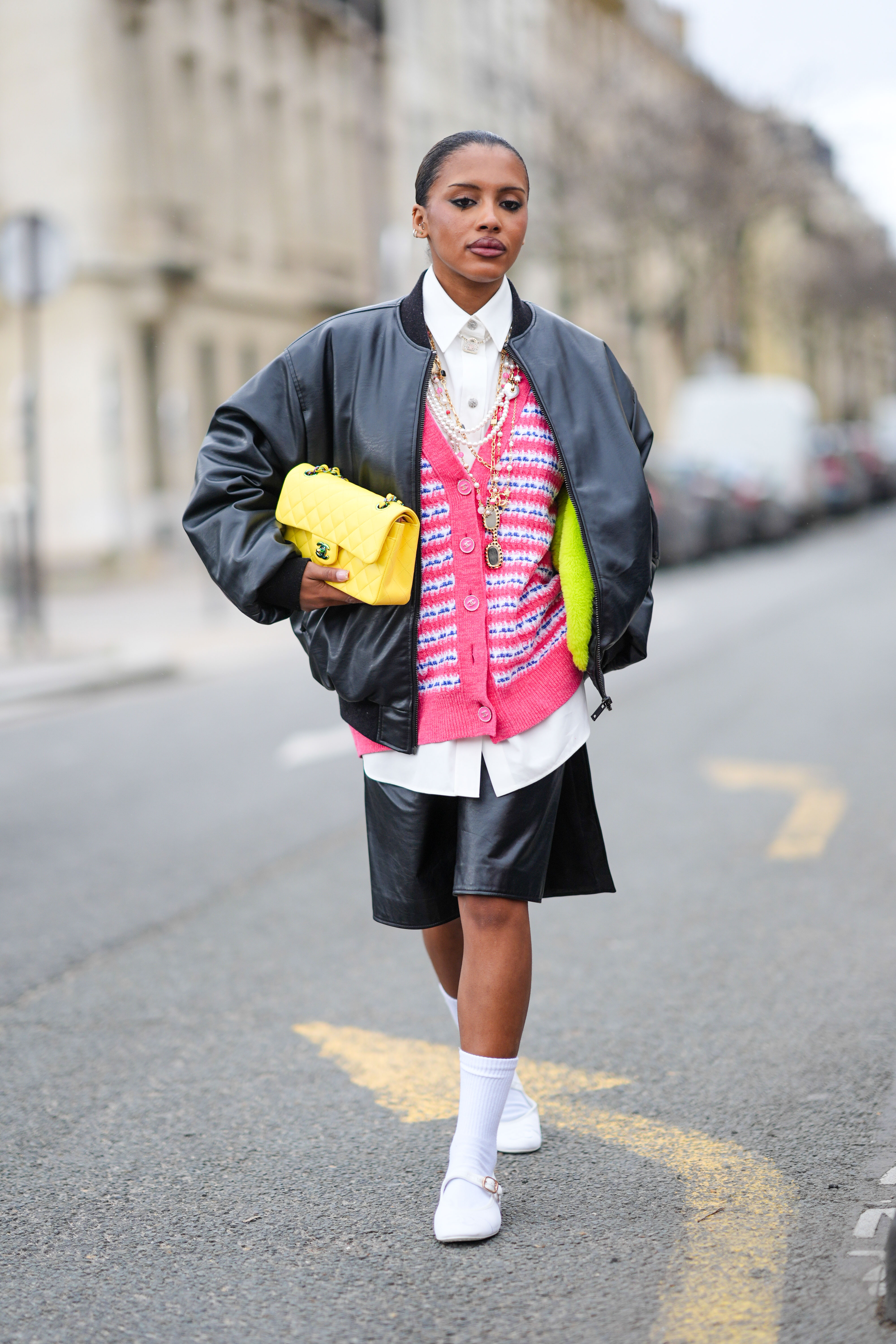 A guest wears a black bomber jacket , a white shirt, a yellow quilted leather Chanel bag, a pink tweed cardigan, a black leather skirt , white socks, white shoes, outside Chanel Paris Fashion Week 2024
