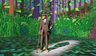 David Hockney standing in front of his digital painting of a forest, projected onto the wallks of a gallery