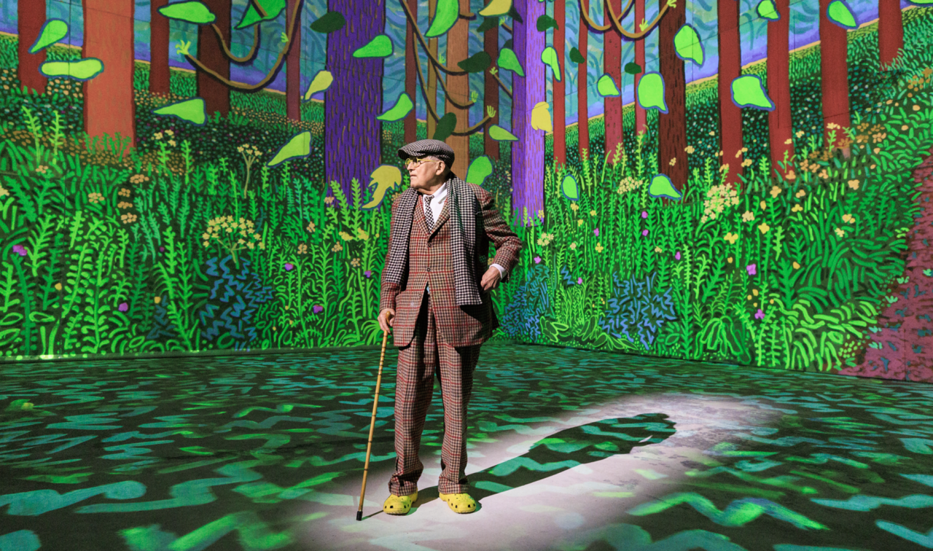 David Hockney standing in front of his digital painting of a forest, projected onto the wallks of a gallery