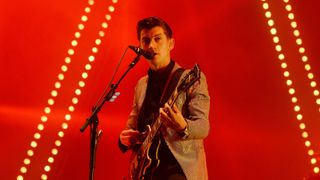 Lead singer of the Arctic Monkeys playing guitar against the red background of the Pyramid Stage at the Glastonbury Festival.