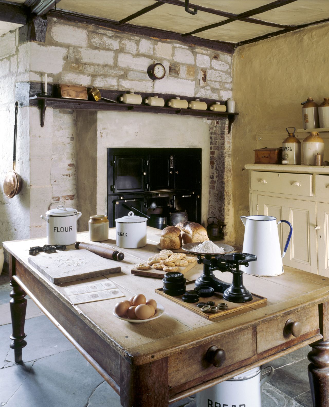 Room view of the Kitchen showing a large table, fireplace and range. The Kitchen range was inserted in the 1930's. The kitchen implements date from the late nineteenth century to the 1950's.