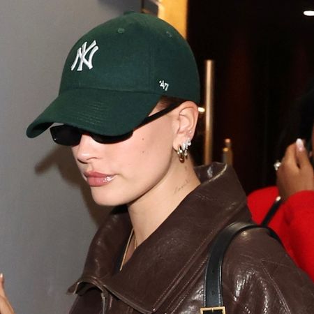 Hailey Bieber wears a green yankees hat, saint lauren sunglasses, saint laurent leather jacket, and saint laurent black bag.