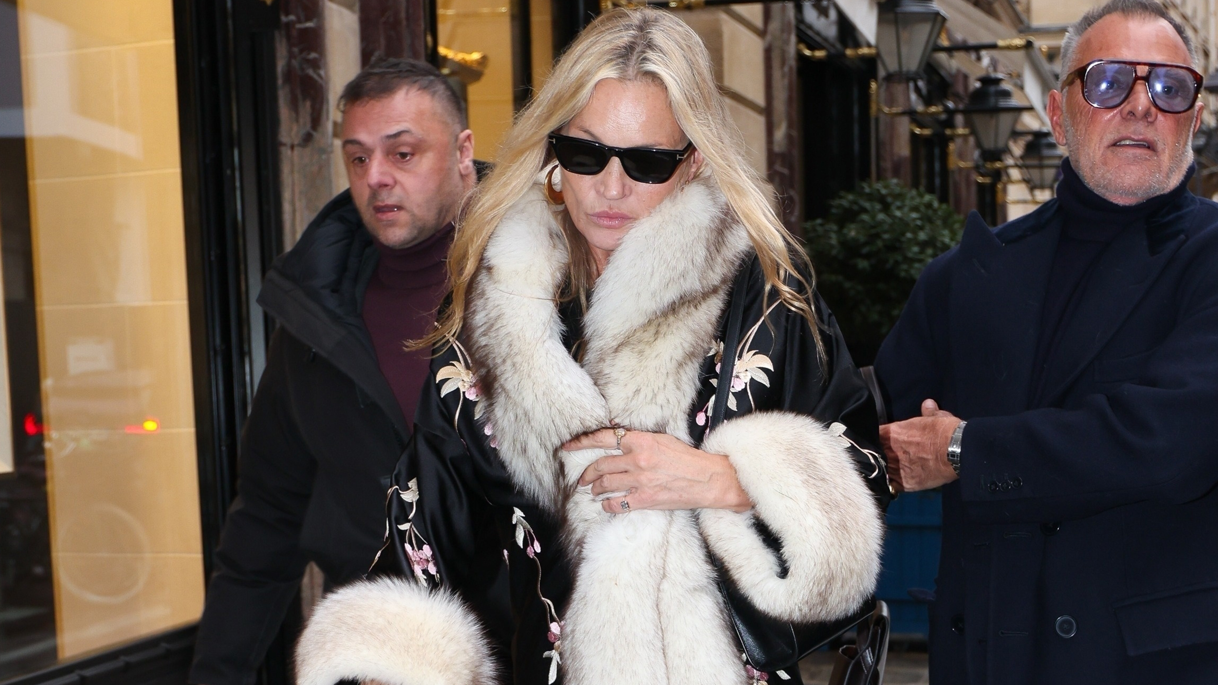 Kate Moss wears black sunglasses with a fur-trimmed coat.
