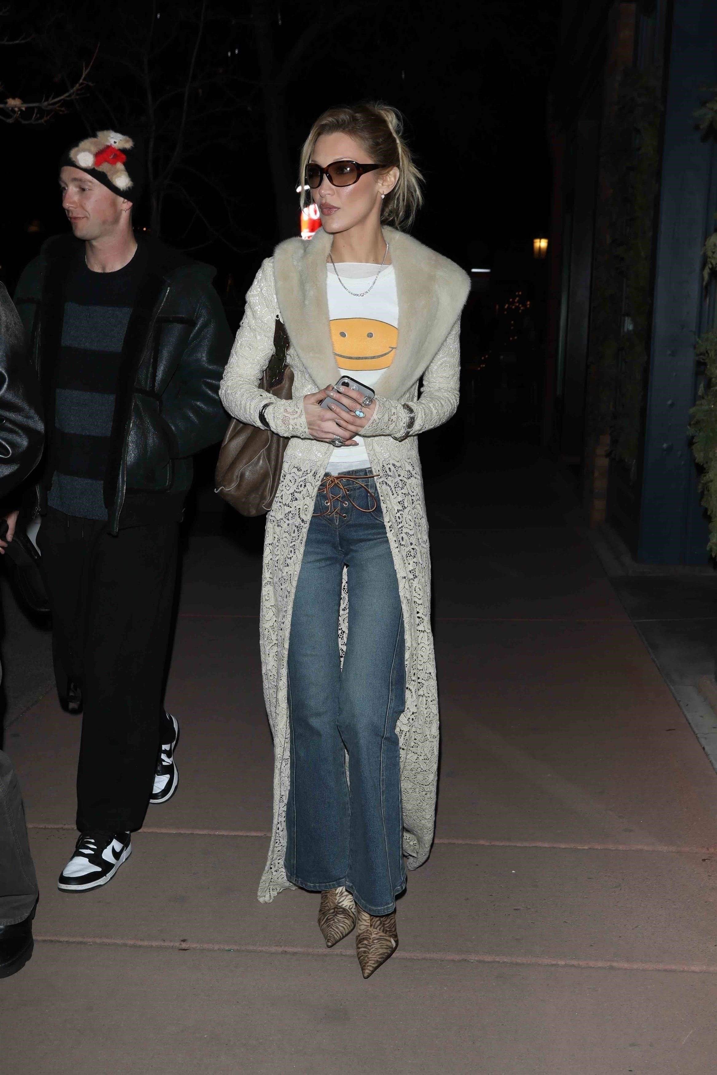 Bella Hadid walks along the pavement outside wearing flared jeans with pointed-toe boots and a lace long-line jacket.