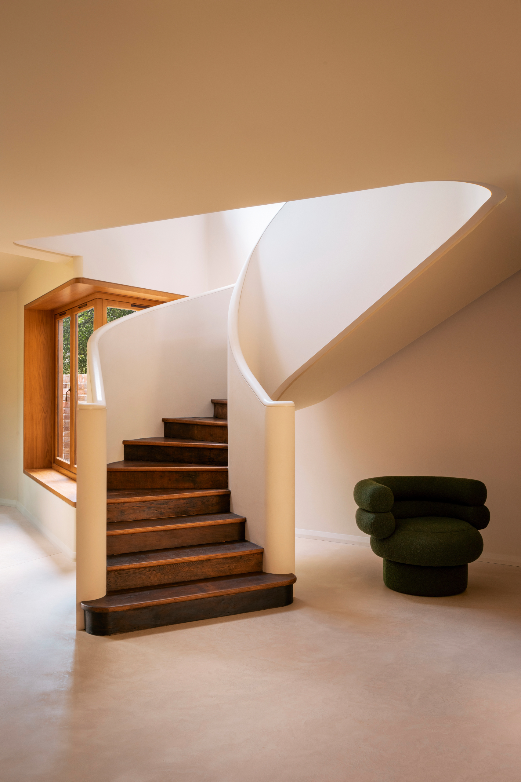 Curved enclosed staircase with cream walls and dark wood steps, leading to a room with microcement floor and a sculptural dark green velvet accent chair