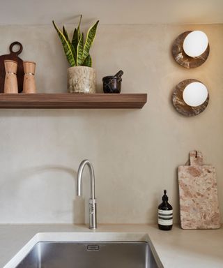 A minimalist kitchen corner with a stainless steel sink, curved faucet, floating wooden shelf, warm wall sconces, and earthy decor, including a potted plant.