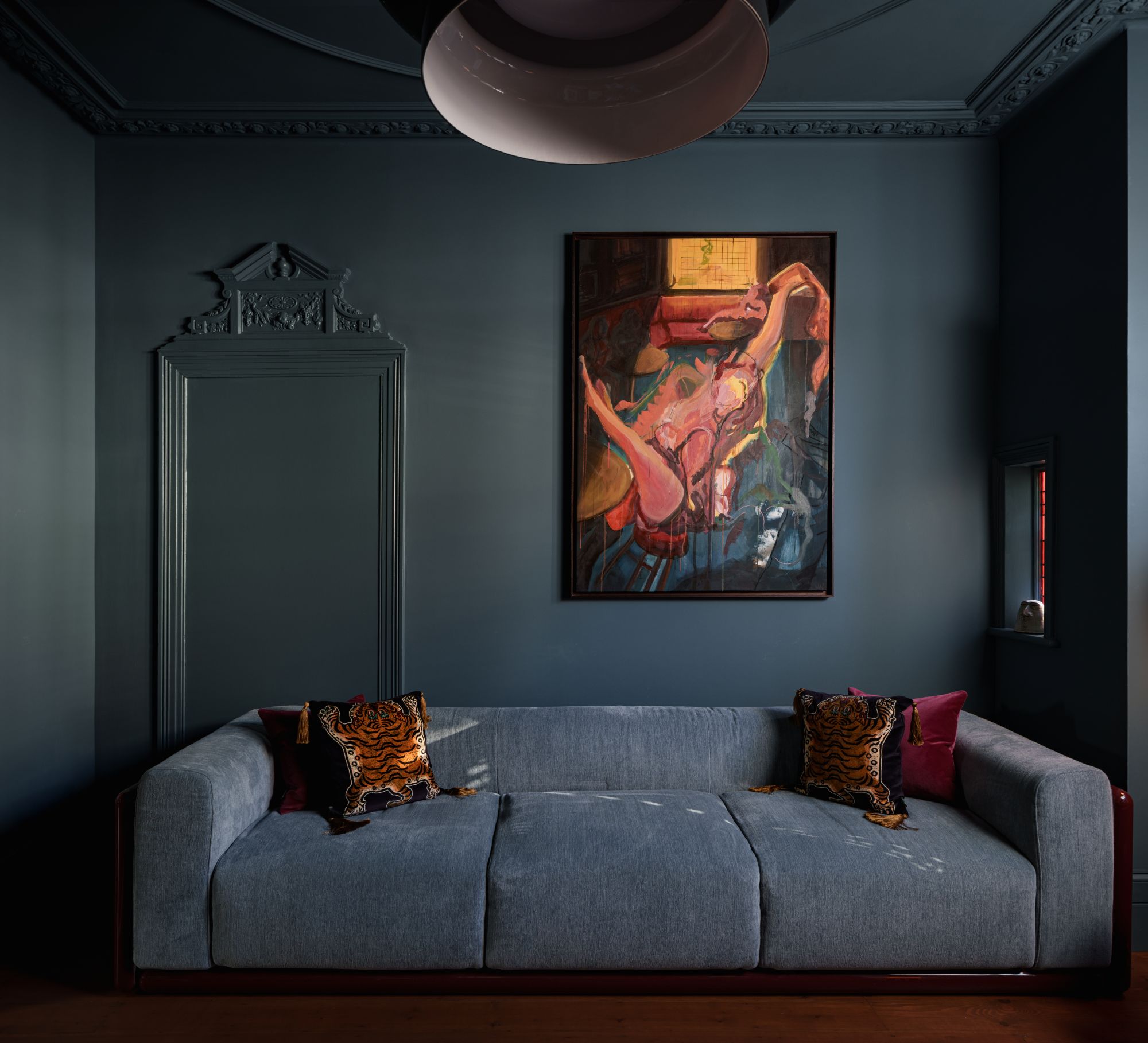 A living room sofa in blue which tones with the teal walls behind and contrasts with the colorful piece of art on the wall and the burgundy and gold-detailed throw pillows