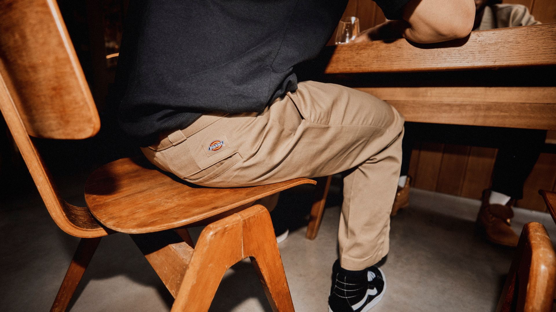 an image of a model wearing the Dickies 247 pants in desert sand whilst sitting down on a wooden chair