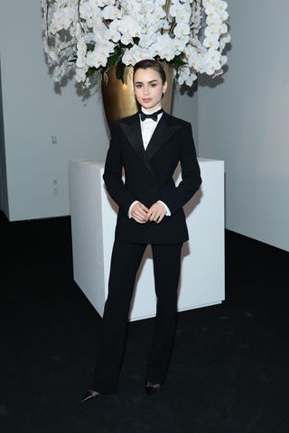 Lily Collins attends the Ralph Lauren Fall 2022 fashion show at Museum of Modern Art in New York City wearing a black fitted tuxedo with high-collar white shirt and black pointed-toe pumps