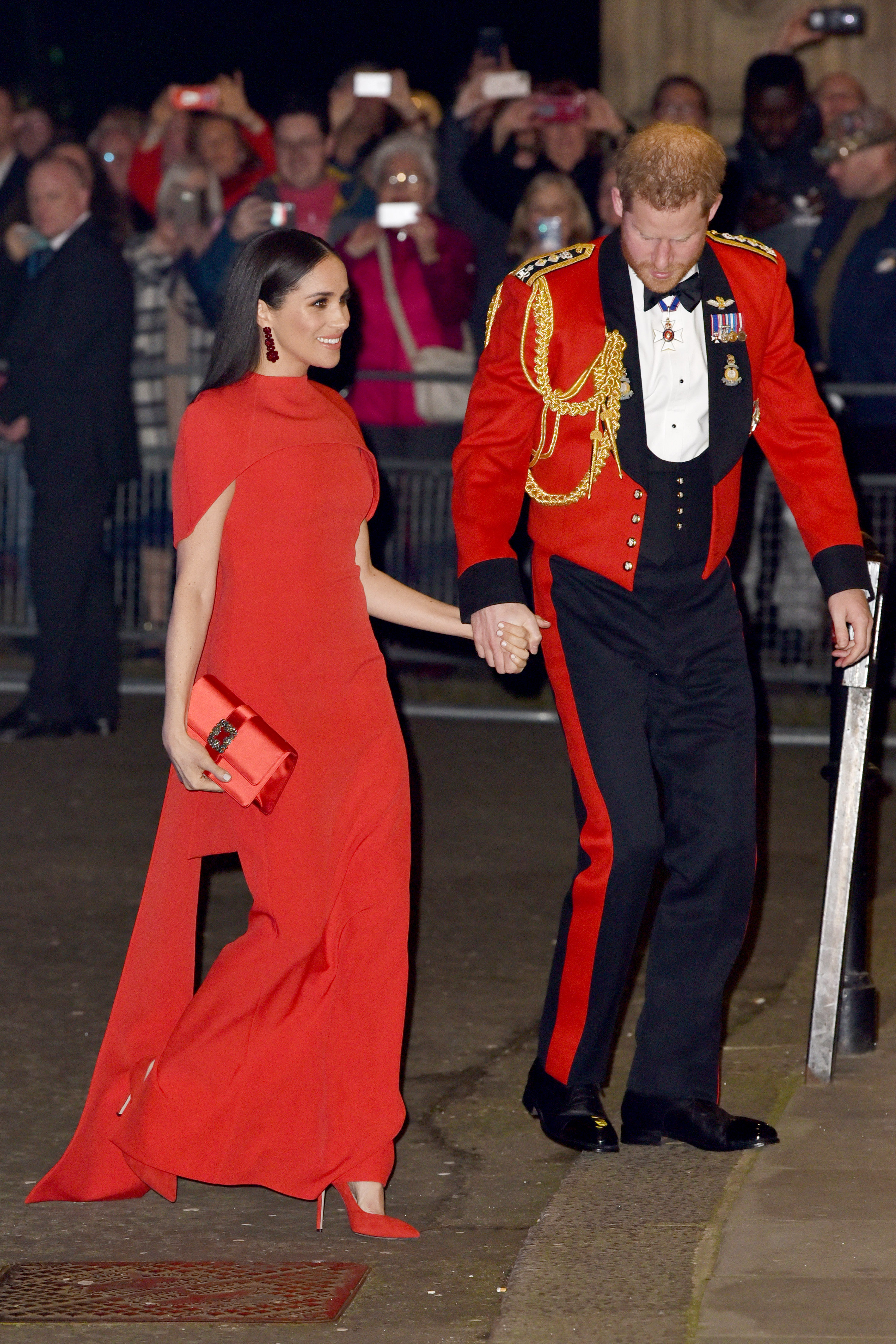 Meghan Markle wearing a red gown and heels holding hands with Prince Harry, wearing a red military coat and black pants