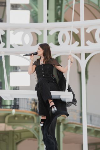 Riley Keough sitzt auf einer Schaukel im Grand Palais und trägt bei der Modenschau Frühjahr/Sommer 2025 ganz in Schwarz Chanel, während er singt 