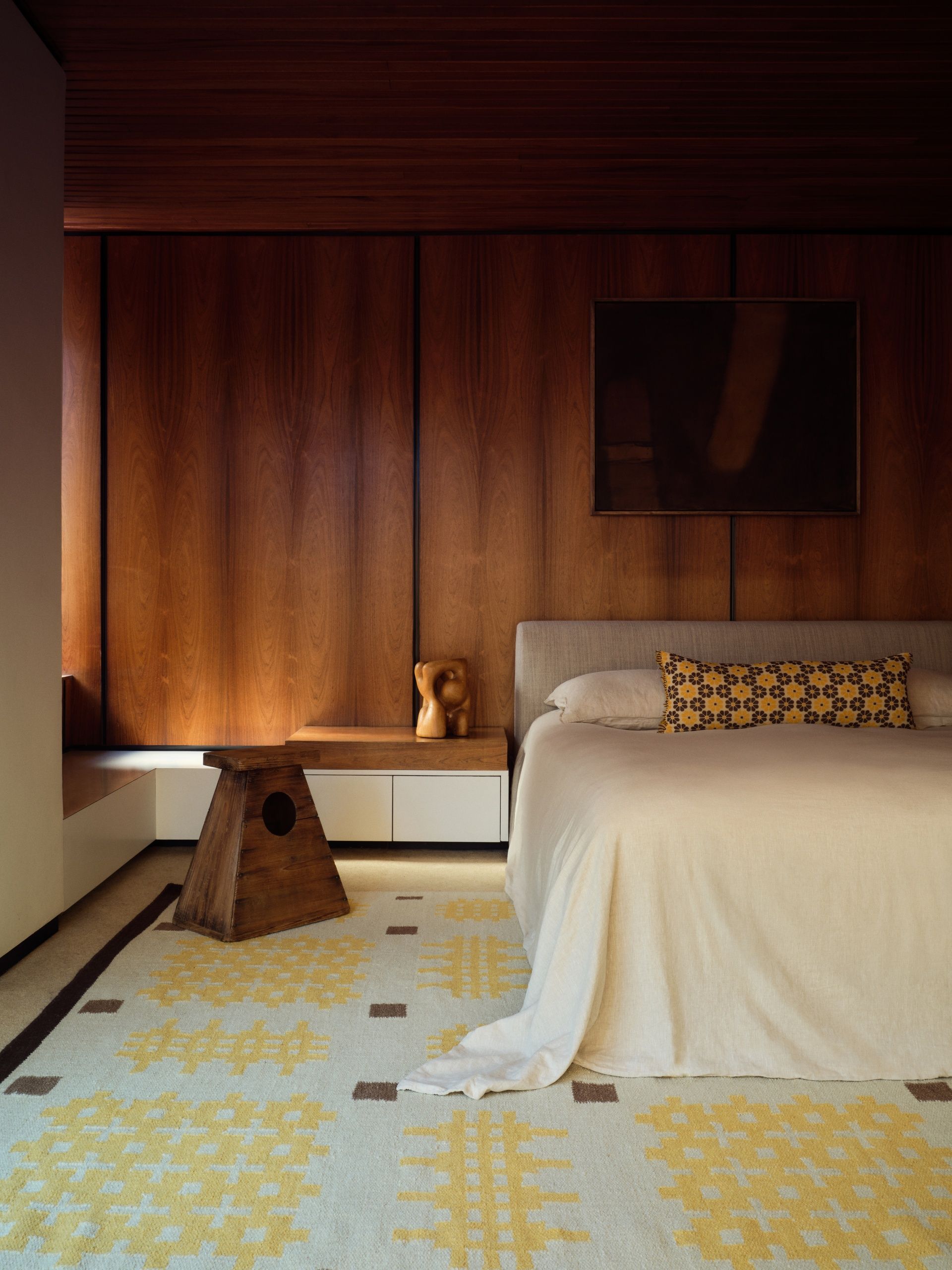 Mid-century style bedroom with wooden walls, a minimalist bed with a yellow and brown bolster pillow and a matching rug.